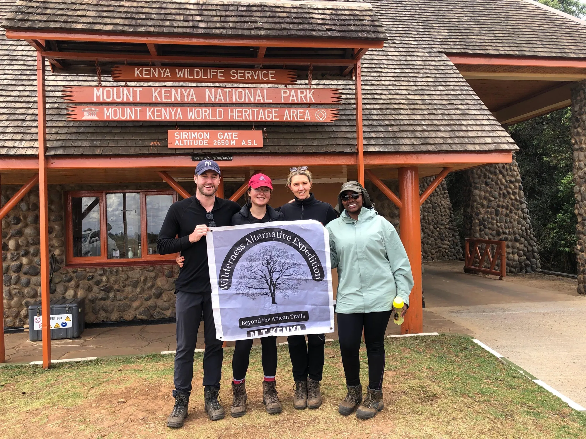 Four guests holding a Wilderness Alternative flag at Sirimon Gate, Mount Kenya